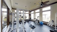 This fitness center in Waxahachie is designed to offer a comprehensive workout experience in a bright and welcoming environment. The space is defined by its generous ceiling height, accented by warm crown molding and fitted with dual ceiling fans that ensure comfortable air circulation throughout. Large, floor-to-ceiling windows line multiple walls, flooding the room with abundant natural light and offering pleasant outdoor views — creating an open, airy atmosphere that enhances the workout experience. The rubber-floored exercise area is well-appointed with a diverse selection of equipment, including a multi-station cable and weight machine branded with Inflight Fitness, a rowing machine, a stationary elliptical, a cable row station, and a weight bench. A stability ball and additional free weights round out the available fitness tools. Recessed lighting supplements the natural light, and a mounted flat-screen television provides entertainment during workouts. The neutral, beige wall tones and clean architectural finishes lend the space a polished yet functional aesthetic. Overall, this fitness center strikes an effective balance between professional gym functionality and a comfortable, well-designed interior setting.