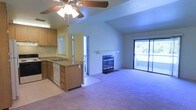 This single-floor residence offers an open and airy layout that seamlessly connects the living and kitchen areas. The living room is a standout space, featuring vaulted ceilings that enhance the sense of volume and openness. A ceiling fan with a light fixture is centrally mounted, providing both airflow and illumination. A fireplace serves as a focal point along the main wall, adding warmth and character to the space. Plush carpeting extends throughout the living area, contributing to a comfortable and inviting atmosphere. A large sliding glass door floods the room with natural light and provides direct access to a balcony, offering views of the surrounding greenery outside.

The kitchen is positioned adjacent to the living area in an open-concept arrangement, featuring warm wood-toned cabinetry that provides generous storage space. The kitchen includes a built-in dishwasher, a sink, and countertop workspace, making it both functional and practical. A window above the sink allows for additional natural light. The transition between the kitchen and living area is smooth, making the overall layout well-suited for everyday living and entertaining. The textured ceiling throughout adds a classic finishing detail to the space.