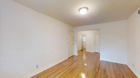 This single-floor space presents a straightforward and functional layout characterized by its clean, minimalist aesthetic. The main living area is finished with polished hardwood floors in a warm honey tone, which reflect light effectively and lend a bright, welcoming feel to the room. White walls throughout contribute to an open and airy atmosphere, while flush-mounted ceiling lighting provides consistent illumination.

From the main living area, a doorway leads to an adjoining hallway or secondary space, where a window allows natural light to filter through, brightening the interior. Two white-painted doors are visible beyond the main room, suggesting additional rooms or utility spaces such as a bathroom or closet, contributing to the overall functional layout of the unit.

White baseboards frame the flooring throughout, adding a subtle architectural detail that complements the clean, neutral palette of the space. The single-story design ensures easy accessibility, and the consistent use of hardwood flooring and white finishes throughout creates a cohesive and straightforward interior that serves as a versatile, move-in-ready canvas.