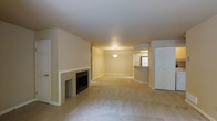 This single-floor residence offers a thoughtfully arranged open layout that connects the main living area to the kitchen, creating a cohesive and functional flow throughout the space. The living room is carpeted in a neutral tone, lending a warm and understated ambiance to the room. A classic brick fireplace with a white-painted surround serves as the focal point of the living area, adding both character and a sense of coziness to the space. The open floor plan transitions seamlessly into a dining or secondary living area, illuminated by a central pendant light fixture that draws the eye and adds a touch of elegance. Adjacent to this area, a breakfast bar connects the living space to the kitchen, which features white cabinetry and appliances, providing a clean and practical culinary workspace. A white paneled door provides access to an additional room or storage area, contributing to the home's overall functionality. The walls and ceilings are finished in a consistent neutral palette throughout, giving the space a unified and inviting atmosphere well-suited for comfortable everyday living.