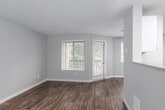This single-floor residence offers a well-organized layout designed for comfortable everyday living. The open living area is a standout feature, showcasing rich, dark hardwood-style flooring that contrasts beautifully with the neutral gray and beige wall tones throughout. A textured popcorn ceiling adds classic character to the space, while multiple windows fitted with horizontal blinds flood the room with generous natural light and offer views of surrounding greenery. A ceiling fan provides added comfort and airflow to the living area. A full-view patio door connects the interior to an outdoor space, enhancing the sense of openness and providing easy access to fresh air. The kitchen is thoughtfully designed with white cabinetry, stainless steel appliances including a refrigerator and range, and lighter wood-style flooring that distinguishes it from the main living zone. Recessed panel lighting ensures the kitchen is well-lit and functional. The bathroom includes a full-sized soaking tub with a shower combo, finished with clean, light-toned surrounds and chrome fixtures. The bedroom, visible in the floor plan, is carpeted and offers a quiet retreat. The overall ambiance of the space is clean, neutral, and move-in ready, making it a practical and inviting home.