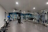 This fitness center offers a comprehensive workout environment within a single, open-plan floor. The space is defined by bold teal accent walls contrasted against neutral white and gray tones, creating an energetic yet focused atmosphere. Durable rubber flooring spans the entire area, providing a practical surface suited for a wide range of exercises.

The cardio zone is well-stocked with multiple treadmills and elliptical machines, each equipped with individual monitors for entertainment or workout tracking. Complementing the cardio section, the strength training area includes a multi-station cable machine, Cybex equipment, a fully stocked dumbbell rack with a tiered weight selection, and additional resistance machines along the walls. A dedicated spin bike and rowing machine round out the equipment offerings.

Full-length mirrors line one wall, visually expanding the space while allowing users to monitor their form. Recessed lighting and ceiling fans maintain a bright, well-ventilated environment throughout. Large windows along one side of the room invite natural light and offer views of the surrounding greenery, adding a refreshing outdoor connection to the workout experience. A small built-in counter area near the entry provides a convenient station for check-ins or personal belongings. Fitness center rules are posted near the entrance, reinforcing the professional and organized character of the facility.
