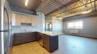 This single-floor space showcases a distinctive industrial aesthetic defined by soaring exposed steel truss ceilings that lend the interior a warehouse-inspired character. The open-concept main area flows seamlessly from the kitchen into the living space, with polished concrete floors reinforcing the modern, utilitarian design throughout.

The kitchen is a standout feature, offering two distinct cabinetry styles: light natural wood upper cabinets paired with dark espresso-toned lower cabinets in one configuration, and a teal blue peninsula island with matching lower cabinetry in another. Both layouts include white quartz-style countertops, stainless steel appliances—including a refrigerator, range, and built-in microwave—and ample drawer and cabinet storage.

A dedicated bedroom area features carpeted flooring in a subtle patterned tile style and exposed concrete block walls painted in a light neutral tone, creating a quieter, more contained space. A window provides natural light and exterior views of the surrounding neighborhood.

The bathroom is well-appointed with a wide vanity featuring dual-tone cabinetry in blue-gray and warm brown finishes, a undermount sink, light countertop surface, and a large frameless mirror. Overall, the space balances raw architectural elements with contemporary finishes throughout.
