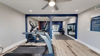 Located at 8546 N 59th Ave in Glendale, Arizona, this fitness center offers a thoughtfully designed single-floor workout space that balances functionality with a clean, modern aesthetic. The interior is defined by light wood-look flooring throughout, crisp white walls accented by bold navy blue architectural columns and trim, and recessed lighting complemented by a ceiling fan that promotes comfortable air circulation during workouts.

The space is divided into distinct workout zones. A cardio area near the entrance features a treadmill, elliptical, and stationary bike, positioned to take advantage of natural light streaming through large glass double doors that open to the exterior. A strength training zone anchors the back of the room, highlighted by an orange power rack with an adjustable bench and a dumbbell rack, all set atop interlocking foam floor tiles for added safety and comfort. Full-length wall mirrors expand the sense of space and allow users to monitor their form during exercise.

A dedicated entry corridor keeps the main workout floor unobstructed, with a posted fitness center rules board and waste bin near the entrance. The large glass-panel doors provide views of the surrounding outdoor area, inviting natural light deep into the space. Overall, the fitness center delivers a well-organized, energetic environment suitable for a full range of training activities.