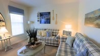 Located on Hayden Dr., this two-story home offers a warm and welcoming atmosphere with a distinct coastal charm that flows throughout its interior. The living area sets the tone for the entire space, showcasing plaid upholstered seating in soft blue and cream tones that evoke a relaxed, beachy sensibility. Nautical accents — including decorative oars, starfish, and seaside-inspired artwork — complement the light, neutral walls and create a cohesive coastal narrative. A sage green oval coffee table anchors the seating arrangement and adds a touch of cottage character, while layered lighting from table lamps casts a soft, cozy glow across the room. Natural light filters in through windows dressed with understated treatments, enhancing the airy feel of the space. The overall ambiance is one of casual comfort and thoughtful design, with curated décor details that feel both personal and purposeful. Spanning two floors, the home provides ample room for living and entertaining, with a layout that balances open communal areas and private retreats. The consistent use of soft, nature-inspired tones and nautical motifs gives the entire home a unified, tranquil character that is both inviting and visually cohesive.
