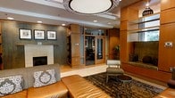 This single-floor space presents a polished and professionally designed interior that exudes a welcoming yet refined atmosphere, characteristic of an upscale hotel lobby or corporate common area. The layout is thoughtfully organized, combining open lounge areas with semi-private enclosed rooms framed by floor-to-ceiling glass panels and warm wood surrounds, offering both transparency and a sense of seclusion.

The lounge area is anchored by rich leather seating and complementary upholstered accent chairs, arranged atop a richly patterned area rug that adds depth and texture to the space. A classic fireplace with a clean stone surround serves as a central focal point on one wall, flanked by framed artwork that adds a curated, gallery-like quality. Built-in shelving with display niches further enhances the sense of craftsmanship throughout.

Overhead, a large decorative drum pendant light fixture commands attention, casting warm ambient light across the space. Recessed lighting supplements the overall illumination, ensuring a bright yet comfortable environment. The color palette draws from warm amber wood tones, neutral stone flooring, and earthy textiles, creating a cohesive and inviting aesthetic. The semi-private glass-enclosed rooms visible in the background appear configured for meetings or focused work, adding functional versatility to the overall space.