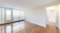 Located at 55 Brownlow Ave in Toronto's Davisville Village neighbourhood, this one-bedroom, one-bathroom unit offers a practical and well-organized layout across a single floor. The living room is a standout space, featuring gleaming parquet hardwood flooring in a classic block pattern and floor-to-ceiling vertical blinds that frame a sliding glass door leading to a private balcony. Natural light fills the room, offering pleasant outdoor views of the surrounding neighbourhood. The open sightline from the living area connects to the kitchen, which is finished with light tile flooring, white cabinetry with modern hardware, a granite-effect countertop, and a full-size refrigerator. The bedroom continues the parquet flooring theme and is brightened by a large window with baseboard heating beneath it, providing a comfortable and airy atmosphere. The bathroom is clean and functional, featuring a full soaker tub with a shower, white subway tile surround, and chrome fixtures. Throughout the unit, neutral wall tones and white trim create a cohesive, understated aesthetic. A hallway connects the main areas efficiently, completing this well-maintained and thoughtfully arranged home in a sought-after Toronto location.