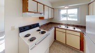 This single-floor 1-bedroom, 1-bathroom residence offers a practical and comfortable layout designed for efficient everyday living. The kitchen is equipped with a white electric coil-burner range, ample upper and lower cabinetry in white and warm wood tones, dark laminate countertops, and a double-basin stainless steel sink positioned beneath a window that brings in natural light. The tile flooring adds durability to this functional workspace. The living area is generously proportioned, featuring wall-to-wall carpeting, neutral walls, and a large window fitted with a wall-mounted air conditioning unit and baseboard heating — providing year-round climate control. A closet with louvered doors adds convenient storage to this main space. The bedroom is a calm, carpeted retreat with clean white walls and ample room for furnishings, bathed in natural light that creates a bright and welcoming atmosphere. The bathroom is finished with tile flooring, a full-sized soaking tub and shower combination with a clean white surround, a dark-toned vanity with granite-style countertop, and brushed nickel fixtures and grab bar accents. The overall design is straightforward and functional, offering a clean, move-in ready living environment.