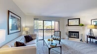 This thoughtfully designed 2-bedroom, 1-bathroom residence offers a comfortable and functional single-floor layout. The inviting living room features plush carpeting, a classic wood-burning fireplace with a white mantel, and large sliding glass doors that flood the space with natural light and open directly onto a private patio with outdoor views of mature trees. The adjoining dining and kitchen area is well-equipped with stainless steel appliances — including a refrigerator, range, microwave, and dishwasher — light wood cabinetry, granite-style countertops, and wood-look flooring. The primary bedroom is a generously sized retreat with carpet flooring, a ceiling fan, corner windows that invite natural light, and access to a walk-in closet with built-in shelving and hanging systems. The second bedroom, equally bright and carpeted, also features a ceiling fan and windows, making it suitable as a home office or guest room. The bathroom is clean and functional, offering a full tub-shower combination and a vanity with ample counter space. Additional highlights include a dedicated laundry room, utility room, hallway, and five closets throughout, providing exceptional storage. The overall ambiance is bright, neutral, and well-organized.