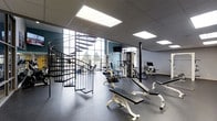 The Haven is a modern, well-appointed fitness facility designed to support a comprehensive workout experience. The space is characterized by its impressive ceiling height, which creates an open, airy atmosphere that makes the room feel expansive and energizing. Large windows along one wall flood the interior with natural light, enhancing the overall sense of openness.

The fitness floor is outfitted with a diverse array of strength-training equipment, including weight benches, cable machines, and multi-use resistance stations, providing options for both targeted and full-body workouts. A dedicated cardio area rounds out the equipment offerings. The flooring consists of durable, dark rubberized material suited for high-activity use.

A striking black spiral staircase serves as both a functional and architectural focal point, adding a sleek, industrial design element to the space. The walls are accented with a muted teal tone, complementing the neutral grey and white palette of the ceiling panels and equipment. Recessed and panel lighting ensures consistent, well-distributed illumination throughout the space, supplementing the natural light. A wall-mounted television provides entertainment during workouts. The overall ambiance is clean, functional, and contemporary, making The Haven's fitness center an inviting environment for residents and users alike.