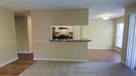 This single-floor space is designed with an open-concept layout that promotes a sense of connectivity between the living and kitchen areas. The kitchen is framed by a distinctive pass-through window cut into a central wall, creating a functional peninsula with dark countertops that serve as both a workspace and a visual dividing element. Through the pass-through opening, the kitchen reveals white cabinetry, a gas range with an overhead ventilation hood, a stainless steel refrigerator, and a single-basin sink — providing a well-equipped cooking environment. The surrounding walls are finished in a warm neutral tone, contributing to a clean and understated aesthetic throughout. The flooring transitions from plush carpeting in the main living area to hardwood-style flooring in the adjacent space, offering both comfort and practicality. Recessed and flush-mount ceiling lighting provides consistent illumination across the space. White baseboards frame the perimeter, adding a crisp finishing detail that complements the overall palette. The layout is straightforward and functional, offering a practical living environment with a cohesive, neutral design scheme.