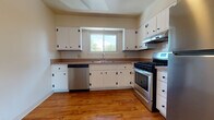 This single-floor residence offers a well-organized layout designed for comfortable everyday living. The kitchen is equipped with crisp white cabinetry accented by black hardware, stainless steel appliances including a gas range and dishwasher, warm hardwood-style flooring, and a window above the sink that brings in natural light and outdoor views. Ample upper and lower cabinet storage makes the space highly functional.

The living area is generously proportioned, featuring neutral-toned carpeting, baseboard heating, and a large sliding glass door with vertical blinds that opens to an outdoor area, flooding the room with natural light. The space flows openly into a secondary area with a hanging chandelier fixture, creating a versatile combined living and dining zone.

The bedrooms are carpeted and bathed in natural light from windows with views of the surrounding neighborhood. One bedroom notably features dual closets, providing substantial storage. Each room is finished with clean, neutral walls and baseboard heating.

The bathroom is finished with classic white subway tile wainscoting, a wood-trimmed vanity with a white countertop and integrated sink, a medicine cabinet above, and tile flooring, offering a clean and timeless aesthetic throughout.