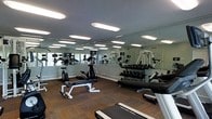 This fitness center offers a fully outfitted workout environment designed to support a wide range of exercise routines. The space is anchored by large floor-to-ceiling mirrors that line the walls, creating a sense of openness and allowing users to monitor their form during exercise. The flooring features durable carpet tile, providing both comfort and stability underfoot. Overhead, a series of recessed panel lights ensure the space is evenly and brightly illuminated throughout.

The facility is equipped with an impressive selection of strength and cardio equipment. A full dumbbell rack holds an extensive range of free weights, while a dedicated weight bench provides a station for targeted strength training. Cardio offerings include stationary bikes and additional aerobic machines, catering to a variety of fitness preferences. Cable and resistance machines round out the strength training options, giving users access to a well-balanced suite of equipment.

The layout is thoughtfully organized to maximize usable floor space, allowing multiple users to work out simultaneously without feeling crowded. The clean, light-toned walls and generous artificial lighting contribute to an energizing and motivating atmosphere, making this fitness center a practical and well-appointed facility for everyday use.