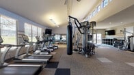 Located at 110 Vanderheck St in San Antonio, TX, this single-story fitness center is thoughtfully designed to support a comprehensive workout experience. The space features soaring ceilings in the main cardio area, where clerestory windows along the upper walls flood the room with natural light, creating an open and energizing atmosphere. The flooring consists of durable carpet tiles in a checkered pattern, providing both comfort and traction across the exercise areas.

The cardio section is lined with a row of True-brand treadmills and elliptical machines positioned alongside large windows that offer outdoor views, complemented by a wall-mounted television for entertainment during workouts. A ceiling-hung heavy punching bag anchors the center of the space, adjacent to a multi-station cable machine ideal for a wide variety of strength exercises.

A dedicated strength training area houses a full dumbbell rack with an organized weight selection, additional cable and resistance machines, and open floor space for functional training. Another section provides isolated strength machines, including leg extension and cable pull-down equipment. Horizontal accent stripes along the neutral-toned walls add subtle visual interest throughout. A sliding glass door at one end offers direct access to the outdoor surroundings, enhancing ventilation and connectivity to the exterior.