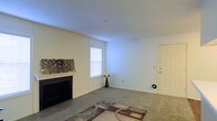 This single-floor space offers a clean and functional layout with a warm, understated aesthetic. The living area serves as the central gathering space, anchored by a classic fireplace with a simple mantel that adds character and a focal point to the room. Decorative accents on the mantel contribute a touch of personality to the otherwise neutral palette.

The flooring throughout is soft wall-to-wall carpeting, providing comfort underfoot and a cozy atmosphere. A patterned area rug further defines the living space, adding visual interest to the room. The walls are finished in a light, neutral tone that reflects natural light effectively, enhancing the sense of openness.

A window fitted with horizontal blinds allows natural light to filter into the space while maintaining privacy. The ceiling features a smooth finish and is equipped with a smoke detector, reflecting attention to safety. A standard interior door provides access to an adjacent room or utility area, contributing to the overall functional flow of the floor plan.

The space presents a straightforward and livable environment, well-suited for those seeking a comfortable, move-in-ready layout with classic residential features throughout.
