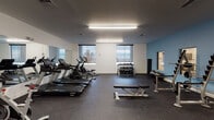 This fitness center offers a comprehensive, single-floor workout facility designed to support a wide range of exercise routines. The space features a durable rubber-speckled flooring throughout, providing both safety and comfort during training sessions. A row of Spirit-brand treadmills and elliptical cross-trainers lines one side of the room, complemented by a full-wall mirror that visually expands the space and allows users to monitor their form. Natural light filters in through multiple windows fitted with roller shades, offering views of the surrounding landscape and creating a bright, energizing atmosphere. Overhead fluorescent lighting ensures the space remains well-lit at all hours. The free weights area includes a multi-tiered dumbbell rack stocked with a comprehensive range of weights, accompanied by an adjustable workout bench. A squat rack with an Olympic barbell and weight plates is also available for strength training. A stationary bike and rowing machine round out the cardio offerings. The walls are finished in a clean white with a calming blue accent wall, contributing to a modern, motivating aesthetic. A dedicated exit door with emergency signage ensures safe egress, and a small waste station keeps the facility tidy. The open layout allows for ample floor space between equipment, accommodating multiple users simultaneously with ease.