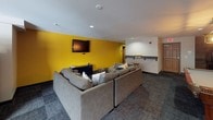 This game room is a well-appointed communal entertainment space designed to offer a variety of leisure activities under one roof. The layout is thoughtfully organized into distinct zones, each catering to different forms of recreation and relaxation.

At the heart of the room sits a classic wooden pool table, illuminated by stylish pendant lighting that casts a warm glow over the playing surface. A wall-mounted television and cue rack nearby add to the functionality of this gaming corner, which is finished with rich wood-paneled accents.

The lounge area is anchored by generous sectional sofas — including both a light neutral sectional and a dark leather option — arranged to encourage conversation and group viewing. A bold yellow accent wall frames a wall-mounted flat-screen TV, creating a vibrant focal point in the seating zone. Accent pillows and a round side table complete the inviting atmosphere.

Near the entrance, a dedicated arcade nook houses multiple classic upright arcade cabinets, including recognizable titles, with wooden bar stools for comfortable play. Natural light filters in through nearby windows, brightening this corner of the room.

Throughout, durable patterned carpet flooring and recessed lighting maintain a cohesive, modern-casual aesthetic. A counter area provides additional surface space, rounding out this dynamic and versatile entertainment room.