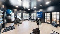 Located at 1900 Switzer Rd in Gulfport, Mississippi, The Bayou District fitness center is a dedicated exercise room designed to meet a variety of workout needs. The space features a bold, modern aesthetic with dramatic matte-black walls and ceiling, contrasted by light hardwood-style flooring that adds warmth and brightness underfoot. Recessed lighting throughout the ceiling provides ample illumination across the entire room.

The exercise room is well-equipped with a diverse selection of fitness equipment, including treadmills, a stationary bike, and a multi-function cable weight machine. A dumbbell rack lines one wall, offering a range of free weights for strength training. Stability balls and foam rollers are neatly stored on wall-mounted shelves, keeping the floor space open and functional. A large wall mirror on one side of the room enhances the sense of space while allowing users to monitor their form during workouts.

Natural light filters in through several windows fitted with white shutters, offering views of the outdoor pool area. The branded "The Bayou District" signage is prominently displayed on the wall, reinforcing the community identity. The space is accessed through classic French-style glass doors as well as a solid panel door, adding a touch of architectural character to this practical and well-appointed fitness facility.