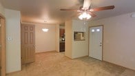 This single-floor residential space offers a practical and comfortable open-concept layout designed for easy everyday living. The main living area is carpeted and generously proportioned, providing a warm and inviting atmosphere. A ceiling fan with an integrated light fixture is centrally mounted overhead, ensuring year-round comfort and ambient lighting throughout the space. The front entry features a solid door with glass panel inserts, allowing natural light to filter into the foyer, which transitions smoothly from tile flooring into the carpeted living area. A pendant light fixture illuminates the dining area, which connects seamlessly to the kitchen. The kitchen is equipped with dark wood cabinetry, built-in appliances including a microwave and refrigerator, and features a pass-through window that opens to the living and dining spaces — enhancing the open feel and making the layout ideal for entertaining or everyday interaction. The neutral color palette throughout, including warm beige walls and light-toned carpet, creates a cohesive and easy-to-furnish environment. Additional doors visible within the unit suggest access to further rooms, adding to the overall functionality of this well-organized single-level home.
