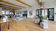 Located at 3612 Beverly Cove Way in Wilmington, NC, this single-story community clubhouse is designed with a contemporary coastal aesthetic that blends comfort and functionality. Wide-plank hardwood flooring runs throughout the entire space, creating a warm and cohesive feel from room to room.

The main lounge is expansive and open, featuring soaring ceilings with exposed wood beams, large black-framed windows that flood the area with natural light, and carefully curated seating arrangements including tufted leather sofas and upholstered accent chairs. Woven pendant lights and original artwork add character and warmth to the social spaces.

A dedicated kitchen and café area showcases shaker-style cabinetry, dark granite countertops, a large central island with bar seating, and stylish cone pendant lighting — ideal for community gatherings or casual use. An adjacent prep area provides additional counter space and a utility sink.

The game room offers a lively retreat with foosball and shuffleboard tables, grey accent walls, a statement chandelier, and direct access to an outdoor pool area through large sliding glass doors and a beverage cooler nearby.

A quiet co-working nook with a marble-topped table and wall sconces positioned beside floor-to-ceiling windows provides a focused workspace with scenic outdoor views, completing this well-rounded and thoughtfully designed community amenity space.