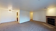 This single-floor space presents a straightforward, functional layout characterized by its open design and neutral finishes throughout. The main living area is generously proportioned, featuring wall-to-wall carpeting in a muted gray tone that provides a soft, understated foundation. White walls and a smooth white ceiling contribute to a bright, clean aesthetic, while standard baseboards frame the perimeter of the room cleanly.

A partial interior wall divides the main living space, creating a natural sense of separation between zones without fully enclosing them. This architectural element adds subtle structure to the open floor plan. Visible beyond this dividing wall is a secondary area, likely a hallway or additional room, illuminated by interior lighting. To the left, an opening leads into what appears to be a smaller room or utility space, partially visible and featuring darker cabinetry — potentially a kitchen or bathroom area.

Electrical outlets are positioned at standard intervals along the walls, and a ceiling vent is visible, indicating a central HVAC system. The overall ambiance is neutral and move-in ready, offering a blank canvas well-suited for a variety of uses and personal styles.