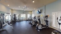 Located at 303 Walkers Mill Cir in Rock Hill, South Carolina, this fitness center offers a well-equipped, single-floor workout facility designed to support a full range of exercise routines. The space is finished with durable rubber flooring throughout, providing a safe and practical surface for all types of training. Soft, neutral-toned walls and recessed lighting create a clean and focused atmosphere, while multiple ceiling fans ensure comfortable air circulation during workouts. Large windows along the walls invite natural light into the space, contributing to an open and energizing environment. The facility is outfitted with a solid selection of cardio machines, including treadmills, elliptical trainers, and a stair climber, as well as multi-station strength equipment and a dedicated free weights area featuring a full dumbbell rack and medicine balls. Wall-mounted mirrors expand the sense of space and allow users to monitor their form. A flat-screen television is mounted on the wall for entertainment during workouts. A keypad-secured entry ensures controlled access to the facility. The overall layout is thoughtfully organized to accommodate multiple users simultaneously, making this fitness center a functional and inviting amenity for the community it serves.