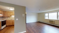 This first-floor unit offers two bedrooms and one bathroom within a well-organized single-level layout. The open living area is a central feature of the space, showcasing warm hardwood-style flooring that extends throughout, creating a cohesive and inviting atmosphere. Neutral wall tones in soft greens and creamy whites keep the space feeling bright and versatile, allowing for a range of furnishing styles.

A window in the living area allows natural light to filter in, enhancing the airy feel of the room. Baseboard heating runs along the perimeter, ensuring consistent warmth throughout the colder months. The living space flows naturally toward the kitchen area, which is partially separated by a dividing wall. The kitchen features warm wood cabinetry, a tiled backsplash, and includes a refrigerator, with a sink positioned near a window for added light while cooking.

The clean white ceilings and simple, functional layout make this unit a practical and comfortable living space. The combination of durable flooring, efficient heating, and a straightforward floor plan provides a solid foundation for everyday living on the first floor.