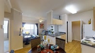 This single-floor residence offers a thoughtfully designed open-concept layout that brings together the dining area, living room, and kitchen in one cohesive, flowing space. The kitchen is outfitted with cream-toned cabinetry, dark countertops, a double sink, and a breakfast bar that serves as a natural divider between the cooking and living areas. Upper cabinets provide ample storage, and the clean white walls contribute to a bright, airy atmosphere. The dining area features a round dark wood table surrounded by woven wicker-style chairs, adding a warm, textural element to the space. Adjacent to the dining area, the living room is generously sized and benefits from large windows that invite abundant natural light, framed by teal curtains that add a pop of color. The living room is furnished with comfortable seating and accented by a ceiling fan for added comfort. Warm hardwood-style flooring runs throughout, unifying the open space. Crown molding along the ceilings adds a refined architectural detail. A hallway visible toward the back of the space suggests additional rooms beyond, rounding out this well-appointed, functional home.