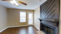 This single-story space showcases a thoughtfully designed open floor plan that seamlessly connects the living and dining areas. The living room serves as the focal point of the home, anchored by a striking dark wood fireplace surround featuring a bold diagonal chevron pattern, a substantial wood mantel, and a raised tile hearth. The fireplace's dramatic dark finish creates a strong visual contrast against the warm beige walls.

Rich, dark hardwood-style flooring runs throughout the main living areas, adding warmth and a cohesive flow between spaces. The living room is well-lit by a large window fitted with horizontal blinds and an exterior door that allows additional natural light to filter in. A ceiling fan with a light fixture provides both air circulation and ambient lighting overhead.

The adjacent dining area is defined by white wainscoting along the lower walls and is illuminated by a decorative chandelier, adding a touch of elegance. A pass-through window connects the dining area to what appears to be the kitchen beyond, promoting an open and communicative layout. The textured ceiling adds subtle architectural character throughout the space.