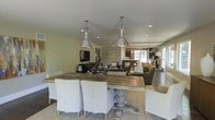 This single-floor space showcases an open-concept layout that seamlessly connects the dining, living, and kitchen areas into one cohesive environment. The dining area anchors the foreground with a substantial rustic wood farmhouse table surrounded by upholstered slipcovered chairs in a classic ticking stripe pattern. Two large industrial-style pendant lights with polished metal shades hang overhead, providing focused illumination while serving as a design focal point.

Recessed lighting throughout the ceiling supplements the natural light that filters in through large windows and a glass door visible toward the rear of the space, lending the interior a bright and airy quality. The walls are finished in a soft, warm neutral tone that complements the earthy, coastal aesthetic carried throughout.

The living area transitions naturally from the dining space, featuring a comfortable upholstered sofa and armchairs arranged around a low coffee table, with a wall-mounted television completing the seating area. Beyond, a kitchen area is partially visible, outfitted with modern appliances. A driftwood centerpiece on the dining table and understated artwork on the walls reinforce the relaxed, coastal-inspired character of the overall space.