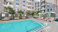 Located along North Coast Highway in Oceanside, California, this outdoor amenity space is designed as a resort-style retreat nestled within a modern multi-story residential building. The centerpiece is a generously sized swimming pool with crystal-clear turquoise water, accessible via a built-in entry rail and flanked by a wide pool deck finished in smooth concrete. Surrounding the pool, tall palm trees and drought-tolerant plantings such as agave and tropical foliage create a lush, coastal atmosphere that complements the sunny Southern California climate. A row of cushioned lounge chairs with mint-green upholstery lines one side of the pool, while slatted wood-and-fabric cabana structures provide shaded retreats for relaxation. On the opposite side, additional lounge seating with colorful umbrellas offers open-air sunbathing options. A separate outdoor living area features terracotta tile flooring and is appointed with a large sectional sofa accented with teal and coral cushions, a slatted wood coffee table, and woven accent chairs — ideal for social gatherings. The space is framed on multiple sides by the building's contemporary grey and white facade, with balconies overlooking the courtyard. Pendant-style outdoor lighting adds ambiance for evening use.