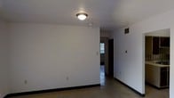 This single-floor space offers a straightforward and functional layout designed for practical living. The main room features smooth white walls, dark baseboards, and terrazzo flooring that adds a classic, durable character underfoot. A flush-mount ceiling light fixture provides overhead illumination, while a ceiling-mounted smoke detector and a visible HVAC vent contribute to the room's utility. The walls include standard electrical outlets and a light switch, reflecting a no-frills, functional design approach. A hallway extends from the main room, leading to additional areas of the space. At the end of the corridor, a window allows natural light to filter through, brightening the interior and offering a glimpse of the outdoor surroundings. The overall ambiance is clean, minimal, and unadorned, making the space a blank canvas well-suited for customization. The consistent use of white throughout the walls and ceiling creates a bright, open feel, while the terrazzo flooring ties the rooms together with a cohesive, timeless aesthetic. This space is ideal for those seeking a simple, move-in-ready environment with a neutral palette and solid foundational features.