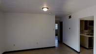 This single-floor space offers a straightforward and functional layout designed for practical living. The main room features smooth white walls, dark baseboards, and terrazzo flooring that adds a classic, durable character underfoot. A flush-mount ceiling light fixture provides overhead illumination, while a ceiling-mounted smoke detector and a visible HVAC vent contribute to the room's utility. The walls include standard electrical outlets and a light switch, reflecting a no-frills, functional design approach. A hallway extends from the main room, leading to additional areas of the space. At the end of the corridor, a window allows natural light to filter through, brightening the interior and offering a glimpse of the outdoor surroundings. The overall ambiance is clean, minimal, and unadorned, making the space a blank canvas well-suited for customization. The consistent use of white throughout the walls and ceiling creates a bright, open feel, while the terrazzo flooring ties the rooms together with a cohesive, timeless aesthetic. This space is ideal for those seeking a simple, move-in-ready environment with a neutral palette and solid foundational features.