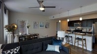 This single-floor residence showcases an open-concept layout that integrates the living, dining, and kitchen areas into one cohesive and functional space. The living room is anchored by a dark upholstered sofa accented with blue and teal throw pillows, complemented by a ceiling fan with integrated lighting overhead. A table lamp with a cylindrical shade adds warm, ambient lighting to the seating area. Framed wall art in a coordinated series adds a curated, contemporary touch to the neutral-toned walls. The dining area sits adjacent to the living space, featuring a wooden table with matching chairs and a simple centerpiece arrangement. Transitioning toward the kitchen, a peninsula counter with bar-height seating serves as a natural divider. The kitchen is equipped with dark cabinetry, stainless steel appliances including a microwave and refrigerator, and pendant lighting that hangs above the counter. Warm wood-tone flooring runs throughout, tying the spaces together cohesively. The overall ambiance is modern and inviting, with a neutral color palette punctuated by coordinated blue accents.