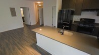 This contemporary single-floor residence offers a thoughtfully designed open-concept layout that seamlessly connects the kitchen and living areas. The kitchen serves as the heart of the space, anchored by a generous central island topped with a warm-toned countertop and fitted with an undermount sink and a matte black faucet. Dark espresso cabinetry lines the upper and lower sections of the kitchen, providing ample storage while lending a sophisticated contrast to the light-colored walls. A black stainless steel refrigerator anchors the kitchen's main wall, adding to the cohesive, modern aesthetic. Rich, dark hardwood-style flooring runs throughout the space, grounding the interior with warmth and visual continuity. The open hallway visible beyond the kitchen leads to additional rooms, each accessible through clean, panel-style doors that maintain the home's understated, modern design language. The neutral wall tones and recessed lighting contribute to a bright, welcoming atmosphere. Overall, the space strikes a balance between functional design and contemporary style, making it a practical yet visually appealing living environment.