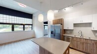 This thoughtfully designed single-floor residence offers a clean, contemporary aesthetic throughout. The open-concept kitchen serves as the heart of the space, featuring a combination of white upper cabinetry and warm wood-toned lower cabinets, complemented by light quartz countertops and a subway tile backsplash. A central island with pendant globe lighting provides additional prep space and serves as a natural focal point. Stainless steel appliances, including a French door refrigerator, dishwasher, microwave, and electric range, complete the functional and stylish kitchen. Light wood-tone luxury vinyl plank flooring runs throughout, tying each area together seamlessly. The living area is open and bright, flowing naturally from the kitchen. The bedroom offers a peaceful retreat with large windows dressed in blackout roller shades, allowing views of the surrounding greenery and outdoor spaces. A well-sized closet with wire shelving provides ample storage. The full bathroom is finished with a warm wood vanity, quartz countertop, an illuminated rectangular mirror, and a full soaking tub with tiled surround. Accessibility features, including a grab bar, are also present. Track lighting and thoughtful fixture choices throughout add to the modern and welcoming character of this residence.