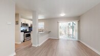 This single-story residence offers a clean, modern aesthetic throughout, unified by light-toned luxury vinyl plank flooring and a neutral gray-and-white color palette. The open-concept layout connects the kitchen directly to the living and dining area, creating a seamless flow ideal for everyday living and entertaining. The kitchen is well-equipped with stainless steel appliances — including a gas range, over-the-range microwave, refrigerator, and dishwasher — complemented by white shaker-style cabinetry, gray quartz countertops, and a single-basin undermount sink. A peninsula with a breakfast bar provides additional counter space and casual seating. The living area is bright and open, drawing natural light through a large sliding glass door that leads to a private outdoor patio. The entry area features a ceiling fan and direct access via a front door with a window. The bedrooms are generously sized, each outfitted with ceiling fans, vertical-blind windows offering views of surrounding greenery, and closet access. The bathroom is tastefully updated with a white shaker vanity, quartz countertop, a framed mirror, and a tiled tub-surround with a built-in niche, maintaining the home's cohesive modern style throughout.