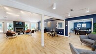 Located at 1747 Capital Circle NE in Tallahassee, Florida, this thoughtfully designed single-floor commercial space offers a generous footprint across an open, flowing layout. The interior is immediately distinguished by bold navy blue accent walls with a textured wood-like finish, set against crisp white trim, vaulted ceilings, and light hardwood flooring that runs throughout the entire space.

The main open area serves as a multifunctional lounge and meeting hub, anchored by a striking herringbone-tiled fireplace with a mounted TV, track lighting overhead, and comfortable seating arrangements. Large windows allow abundant natural light to pour in, with views of a lush outdoor patio area. Adjacent to this is a dedicated dining and collaboration zone, complete with a round table and modern chairs beneath vaulted ceilings adorned with geometric wall art.

A standout feature is the built-in bar area, finished with a dark herringbone countertop, teal tile backsplash, beverage refrigerator, and yellow bar stools — ideal for client entertainment. Private office spaces are also present, equipped with ceiling fans, workstations, and ample room for focused work. A stylish powder room features a wall-mounted matte black sink with modern fixtures. The space also includes a utility/copy room and open shelving for storage, making it fully functional for a professional environment.