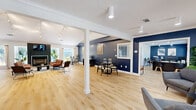Located at 1747 Capital Circle NE in Tallahassee, Florida, this thoughtfully designed single-floor commercial space offers a generous footprint across an open, flowing layout. The interior is immediately distinguished by bold navy blue accent walls with a textured wood-like finish, set against crisp white trim, vaulted ceilings, and light hardwood flooring that runs throughout the entire space.

The main open area serves as a multifunctional lounge and meeting hub, anchored by a striking herringbone-tiled fireplace with a mounted TV, track lighting overhead, and comfortable seating arrangements. Large windows allow abundant natural light to pour in, with views of a lush outdoor patio area. Adjacent to this is a dedicated dining and collaboration zone, complete with a round table and modern chairs beneath vaulted ceilings adorned with geometric wall art.

A standout feature is the built-in bar area, finished with a dark herringbone countertop, teal tile backsplash, beverage refrigerator, and yellow bar stools — ideal for client entertainment. Private office spaces are also present, equipped with ceiling fans, workstations, and ample room for focused work. A stylish powder room features a wall-mounted matte black sink with modern fixtures. The space also includes a utility/copy room and open shelving for storage, making it fully functional for a professional environment.