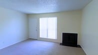 This single-floor space presents a straightforward and functional layout centered around a carpeted living area. The main room features a dark-surround fireplace along one wall, serving as a focal point and providing a practical source of warmth. Adjacent to the fireplace, a large window fitted with vertical blinds allows natural light to fill the room, creating a bright and open atmosphere. Neutral, light-toned walls complement the beige carpeting, contributing to a clean and understated aesthetic throughout. A standard entry door is positioned nearby, offering convenient access to the living area. The textured popcorn ceiling adds a classic residential character to the space. The overall design is simple and uncluttered, making it well-suited for comfortable everyday living. The neutral color palette throughout provides a versatile backdrop, allowing for a variety of furnishing and décor styles.