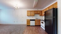 This single-floor residence offers a practical and comfortable layout with clearly defined living spaces. The kitchen is well-equipped, featuring warm honey-toned oak cabinetry — both upper and lower — providing ample storage throughout. Appliances include a white electric range, a black refrigerator, a dishwasher, and a sink with a window-facing position. The kitchen floor is finished in wood-look laminate, which extends into the adjacent open living area. A chandelier overhead adds a touch of classic character to the combined kitchen and living space. The transition from the kitchen into the living area is marked by a shift to neutral wall-to-wall carpeting, which continues through the remainder of the unit.

The living area is generously sized, with neutral white walls, visible ceiling vents, and a door leading to a walk-in storage closet. Natural light filters in through a window, brightening the space. The bedroom is carpeted and includes a closet, a window with blinds, and sufficient space for standard furnishings. The bathroom features wood-look vinyl flooring, a single-sink vanity with oak cabinetry, a full tub and shower combo with grab bar, and a toilet — all finished in clean, neutral tones.