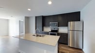 Located at 3443 Park Street in Grove City, Ohio, this single-floor residence showcases a clean, modern design aesthetic throughout. The kitchen is a standout feature, boasting floor-to-ceiling dark espresso cabinetry that provides generous storage while creating a bold, contemporary contrast against the light-toned walls. A sprawling granite-topped island anchors the space, offering ample room for meal preparation and casual dining, complete with an undermount sink and modern faucet. The full suite of stainless steel appliances — including a refrigerator, electric range, and over-the-range microwave — lends a polished, professional feel to the culinary space. A white subway tile backsplash adds a crisp, classic accent behind the range. Recessed lighting illuminates the kitchen evenly, highlighting the quality finishes throughout. Warm wood-toned flooring grounds the space and flows seamlessly into the adjacent areas, complementing the overall neutral color palette. The open layout between the kitchen and living areas enhances the sense of space, making this a well-appointed and functional residence in the heart of Grove City.