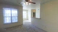 This single-floor residence offers a comfortable and functional layout centered around an open-concept living and dining area. The living space is finished with wall-to-wall carpeting and neutral-toned walls, creating a warm and inviting atmosphere. A ceiling fan with a built-in light fixture is centrally mounted, providing both air circulation and ambient lighting.

Natural light fills the space through multiple windows fitted with horizontal blinds, offering views of the surrounding greenery. The dining area transitions seamlessly from the living room and is illuminated by a pendant chandelier, enhancing the sense of flow between spaces.

A defining architectural feature is the half-wall pass-through that connects the living and dining areas to the kitchen, topped with a dark countertop overhang that serves as a breakfast bar. The kitchen is partially visible beyond the pass-through, where a full-size refrigerator can be seen, indicating a well-equipped cooking space.

Architectural details such as the column-like wall transitions and the open sightlines throughout contribute to the spacious feel of the interior. Overall, the space presents a clean, move-in-ready environment with a straightforward and practical design.