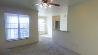 This single-floor residence offers a comfortable and functional layout centered around an open-concept living and dining area. The living space is finished with wall-to-wall carpeting and neutral-toned walls, creating a warm and inviting atmosphere. A ceiling fan with a built-in light fixture is centrally mounted, providing both air circulation and ambient lighting.

Natural light fills the space through multiple windows fitted with horizontal blinds, offering views of the surrounding greenery. The dining area transitions seamlessly from the living room and is illuminated by a pendant chandelier, enhancing the sense of flow between spaces.

A defining architectural feature is the half-wall pass-through that connects the living and dining areas to the kitchen, topped with a dark countertop overhang that serves as a breakfast bar. The kitchen is partially visible beyond the pass-through, where a full-size refrigerator can be seen, indicating a well-equipped cooking space.

Architectural details such as the column-like wall transitions and the open sightlines throughout contribute to the spacious feel of the interior. Overall, the space presents a clean, move-in-ready environment with a straightforward and practical design.