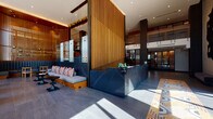 This expansive lobby is a masterfully designed space that blends contemporary architecture with bold, eclectic interiors. Upon entry, visitors are greeted by a striking dark marble reception desk positioned beneath towering vertical wood slat screens that serve as dramatic architectural dividers throughout the space. The double-height ceilings amplify the sense of openness, while floor-to-ceiling glass walls along the perimeter flood the interior with natural light and frame views of the surrounding urban landscape.

The main lounge area is anchored by a sculptural chandelier of cascading metallic branches, suspended above a curated arrangement of curved sage-green seating, vibrant red accent chairs, and dark wood tables arranged atop a graphic navy patterned rug. Teal banquette seating with dark marble tables offers an intimate setting framed by warm wood slat partitions.

A glazed café area features geometric-patterned tile flooring in slate blue and white, complemented by warm wood furnishings and panoramic city views. Secondary corridors showcase textured stone wall cladding and large-format tile floors, leading to glass-enclosed meeting or lounge spaces. A utility alcove features richly tiled amber walls and bold geometric floor patterns. Throughout, the design balances warmth, texture, and contemporary elegance.