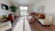 This single-floor, two-bedroom, one-bathroom residence in Monroe, Michigan offers a well-organized layout designed for comfortable everyday living. The living room serves as the heart of the home, featuring wall-to-wall carpeting, a wall-mounted television, and a sliding glass door that opens to a private balcony, flooding the space with natural light and providing outdoor views. The open sightlines create a welcoming and spacious atmosphere throughout.

The galley-style kitchen is efficiently arranged with ample upper and lower cabinetry featuring wood-trimmed accents, a double sink, white appliances, and tile flooring. Adjacent to the kitchen, the dining area accommodates a full table and chairs beneath a multi-bulb chandelier, with a pass-through counter connecting the two spaces.

Both bedrooms are carpeted and well-lit by large windows. One bedroom features dual reach-in closets, offering notable storage capacity, while the second bedroom is bright and serene with sheer curtains enhancing the natural light. The full bathroom includes a single-sink vanity with ample counter space, tile flooring, and a shower-tub combination. Baseboard heating is present throughout the unit, contributing to consistent, efficient climate control.