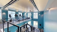 This two-story fitness center offers a well-equipped and visually striking workout environment defined by bold teal walls, white shiplap ceilings, and an abundance of natural light streaming through large multi-pane windows on both levels. The double-height open design creates a spacious, airy atmosphere, enhanced by an oversized ceiling fan that promotes air circulation throughout the facility.

The upper level serves as the primary cardio zone, featuring a row of treadmills and elliptical machines positioned along the perimeter, allowing users to enjoy outdoor views while exercising. A sleek cable railing system lines the mezzanine, providing an open sightline to the floor below while maintaining safety and a modern aesthetic.

The lower level is dedicated to strength and functional training, housing a Life Fitness multi-station cable machine, rowing machines, free weight equipment, and additional cardio bikes. The flooring transitions from carpet in the strength area to tile, helping delineate the training zones. Recessed lighting and wall-mounted accent lights complement the natural light, ensuring the space remains bright and inviting throughout the day. Decorative artwork adds a finishing touch to the energetic, modern environment.