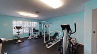 This fitness center is a dedicated, single-floor exercise space designed to support a comprehensive workout routine. The room is finished with vibrant teal-painted walls that create an energetic atmosphere, complemented by durable dark rubber flooring ideal for high-impact activity and equipment placement. Overhead fluorescent lighting ensures the space is well-lit throughout, while large windows fitted with horizontal blinds allow natural light to filter in and provide views of the surrounding outdoor greenery.

The facility is equipped with a well-rounded selection of machines to accommodate both cardio and strength training. Cardio equipment includes a treadmill, a True-brand elliptical trainer, and a stationary cycle, offering multiple options for aerobic exercise. For strength training, the room features Paramount and other multi-station cable machines along with an adjustable weight bench, enabling a range of resistance exercises. A wall-mounted flat-screen television adds an entertainment element to workouts.

Additional amenities include a hand sanitizer dispenser, a waste bin, and a small utility table for personal items. A textured ceiling and clean white baseboards complete the functional yet well-maintained interior. The layout is thoughtfully arranged to maximize usable floor space while keeping equipment accessible, making this fitness center a practical and inviting environment for regular exercise.