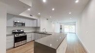 This contemporary single-floor residence showcases a clean, modern aesthetic throughout its thoughtfully designed layout. The open-concept kitchen and living area serves as the heart of the space, featuring a generous quartz-topped island that provides ample prep and gathering space. The kitchen is outfitted with flat-front grey cabinetry extending to the ceiling, stainless steel appliances including a gas range, microwave, and French door refrigerator, complemented by a tile backsplash and a deep undermount sink. Recessed lighting illuminates the space throughout, enhancing its bright, contemporary feel. Warm wood-look flooring runs continuously through the living areas and bedrooms, adding a natural warmth that contrasts elegantly with the crisp white walls. The living area is open and airy, leading directly to an outdoor space through a full-width sliding glass door that floods the interior with natural light. The bedroom features mirrored sliding closet doors and access to an adjoining bathroom. The bathrooms are finished with large-format tile flooring, stone-topped vanities with undermount sinks, and a glass-enclosed walk-in shower, delivering a polished, spa-like experience. Built-in shelving and thoughtful storage solutions add to the overall functionality of this well-appointed space.