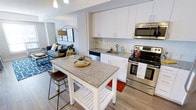 This single-floor residence showcases a thoughtfully designed open-concept layout that seamlessly connects the kitchen and living area, creating an inviting and functional living space. The kitchen is a standout feature, equipped with stainless steel appliances including a range, microwave, and dishwasher. Crisp white cabinetry extends to the upper walls, providing ample storage, while granite countertops add a polished, contemporary feel. A freestanding kitchen island with a matching granite top and lower shelving offers additional prep space and casual seating with a bar-height stool. A classic white subway tile backsplash completes the clean, modern aesthetic. Light hardwood-style flooring runs throughout, lending warmth and visual continuity between spaces. The adjoining living area is bright and open, benefiting from natural light and a comfortable, spacious feel. The open floor plan promotes easy flow between the kitchen and living spaces, making the unit well-suited for both everyday living and entertaining. The overall ambiance is modern and airy, characterized by a neutral color palette and clean architectural lines.